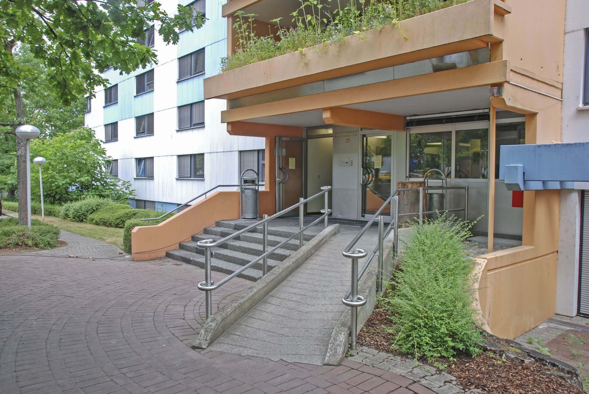 Haupteingang des Wohnheims mit Treppen, Rampe, Handlauf und orangefarbener Fassade.