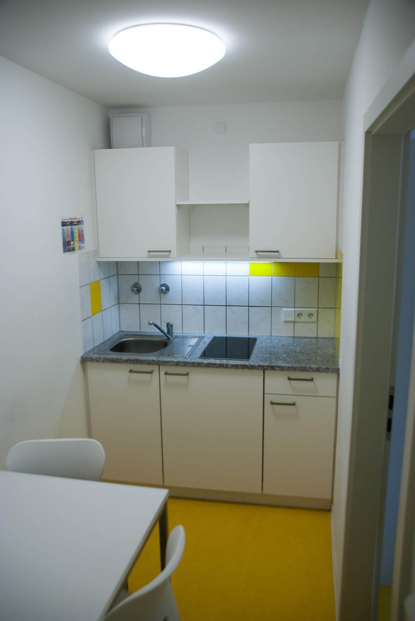 Kompakte Teeküche mit zwei Herdplatten, Spüle und weißen Schränken, mit gelben Akzenten an der Wand.