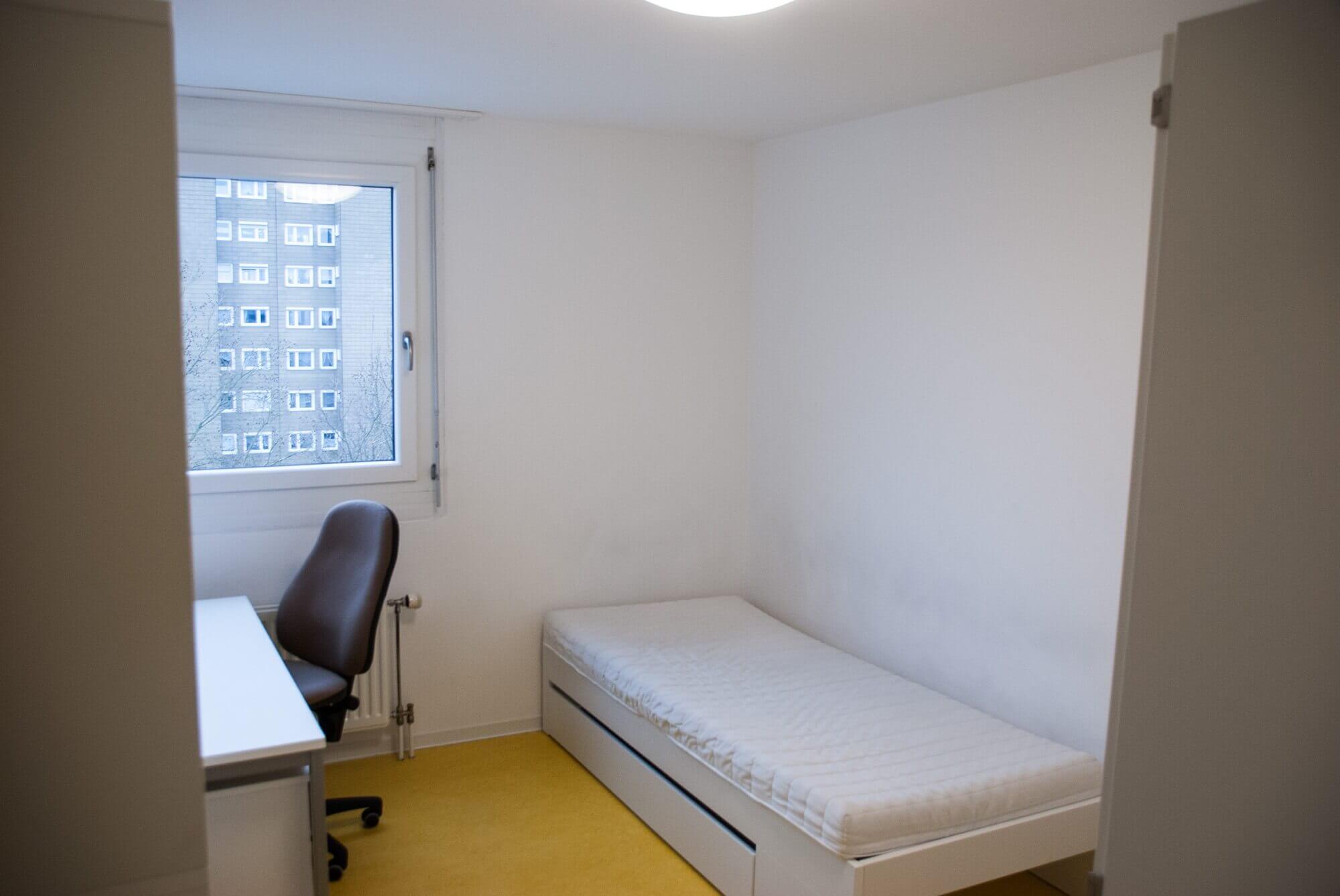 Blick in ein helles Studentenzimmer mit Schreibtisch, Bürostuhl, Bett und Fenster mit Aussicht auf ein gegenüberliegendes Wohngebäude.