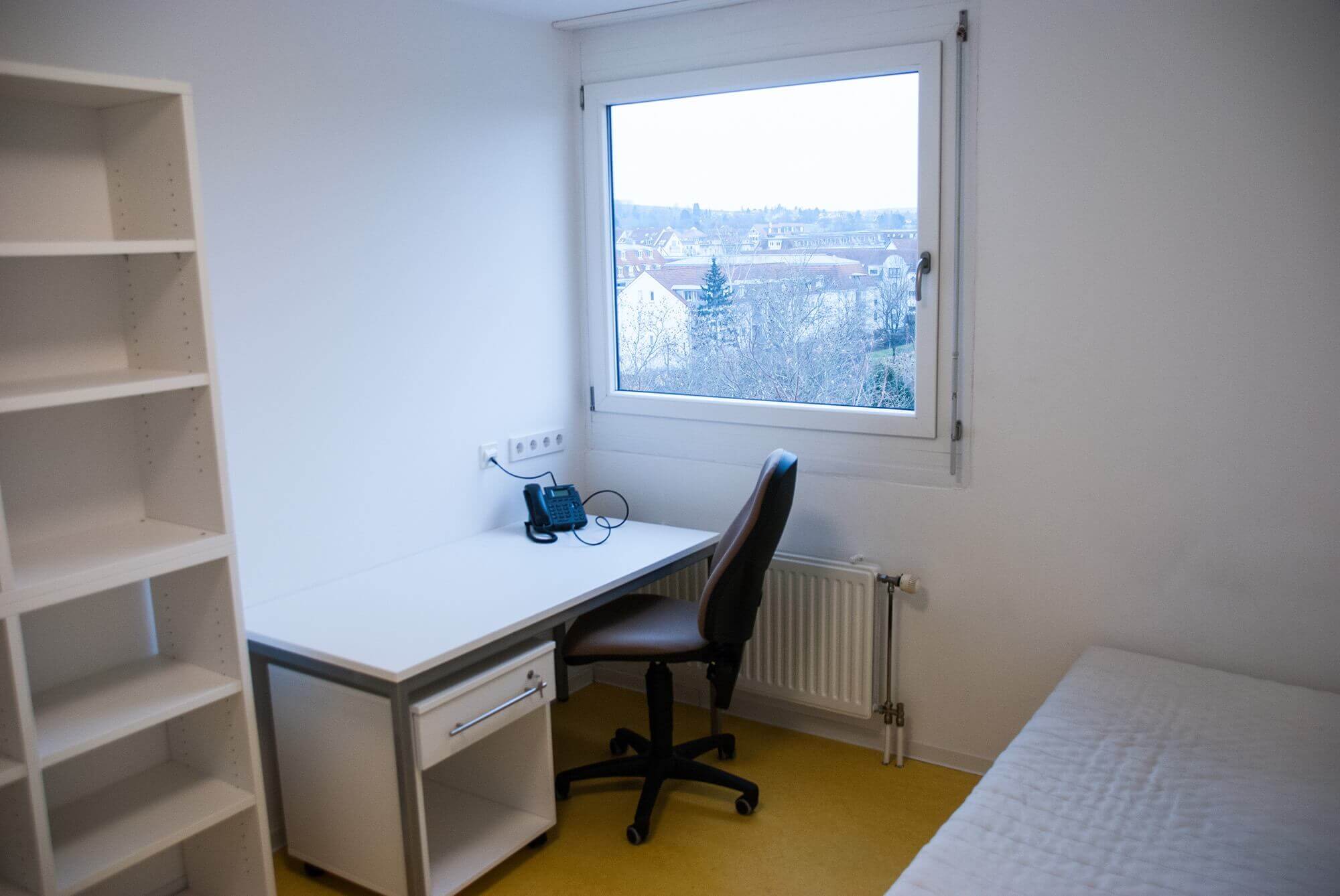 Arbeitsplatz im Studentenzimmer mit großem Fenster, Schreibtisch, Stuhl und einem Regal an der Wand.