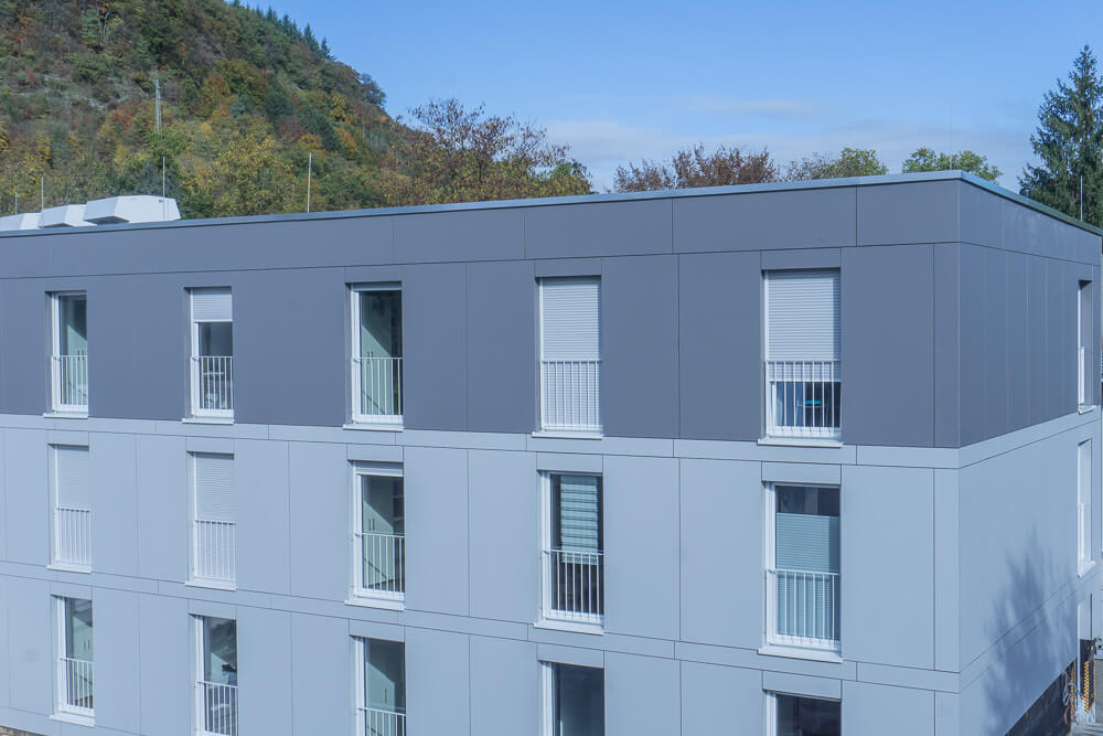 Ein modernes, grau-weißes Apartmentgebäude mit geschlossenen Fensterläden vor der Kulisse grüner, baumbedeckter Hügel unter einem klaren blauen Himmel.