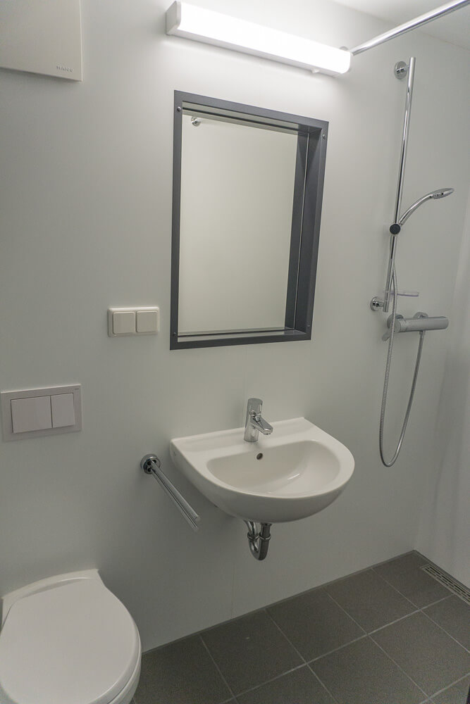 Ein modernes, barrierefreies Badezimmer mit Wandwaschbecken, Spiegel, Dusche mit Handbrause und Toilette. Wände und Armaturen sind weiß, der Boden ist mit grauen Fliesen ausgelegt.