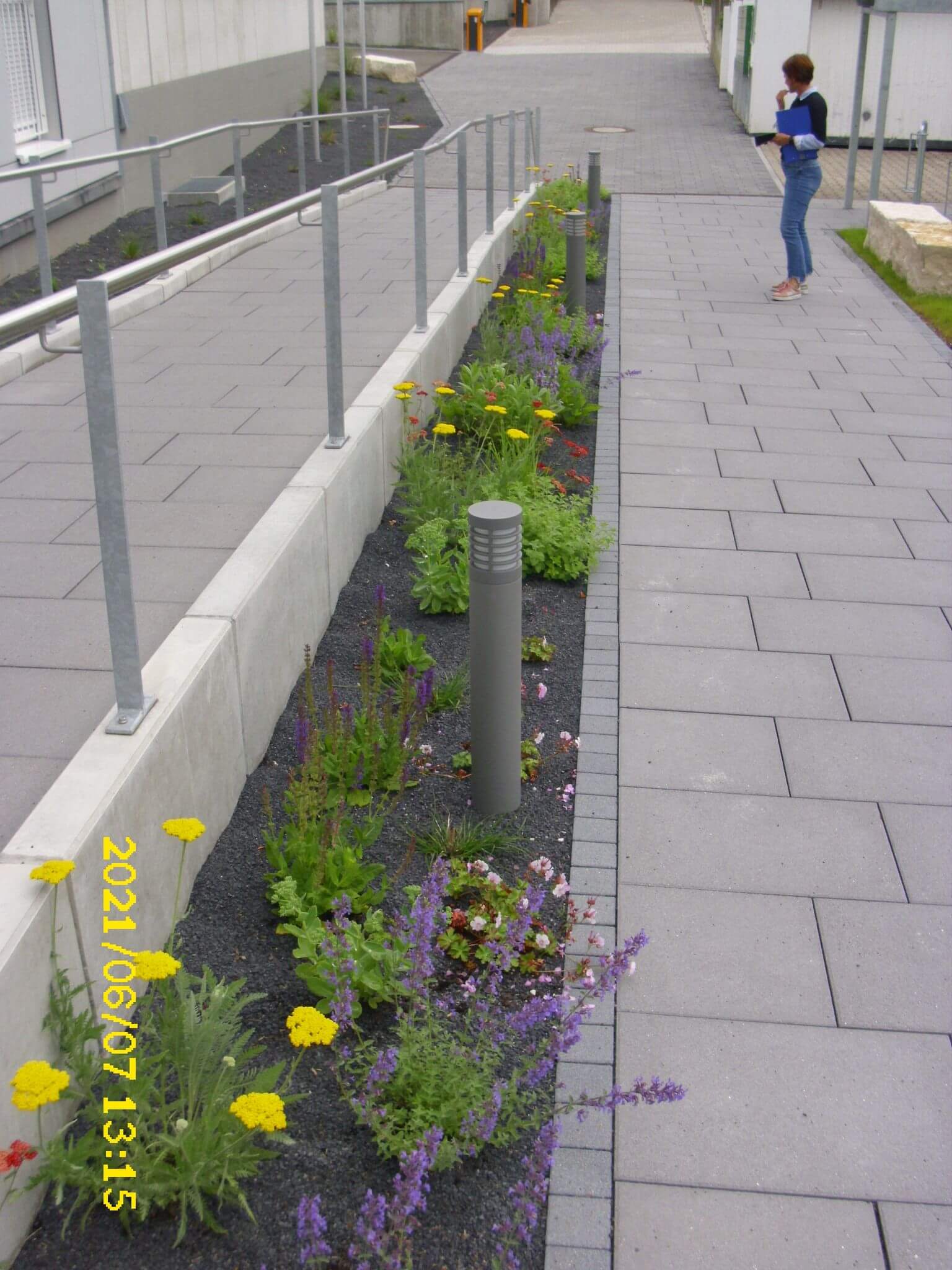 Ein gepflasterter Gehweg mit Geländer verläuft neben einem schmalen Gartenbeet mit bunten Blumen und einem Laternenpfahl. Am anderen Ende steht eine Person und schaut in ein Notizbuch. Der Datumsstempel lautet: 07.06.2021, 13:15 Uhr.