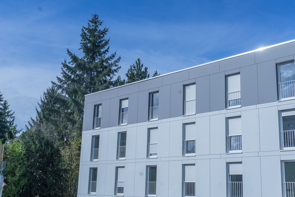 Ein modernes Apartmentgebäude mit hell- und dunkelgrauen Außenpaneelen, mehreren Fenstern mit weißen Fensterläden und kleinen Balkonen vor der Kulisse hoher grüner Bäume und eines klaren blauen Himmels.