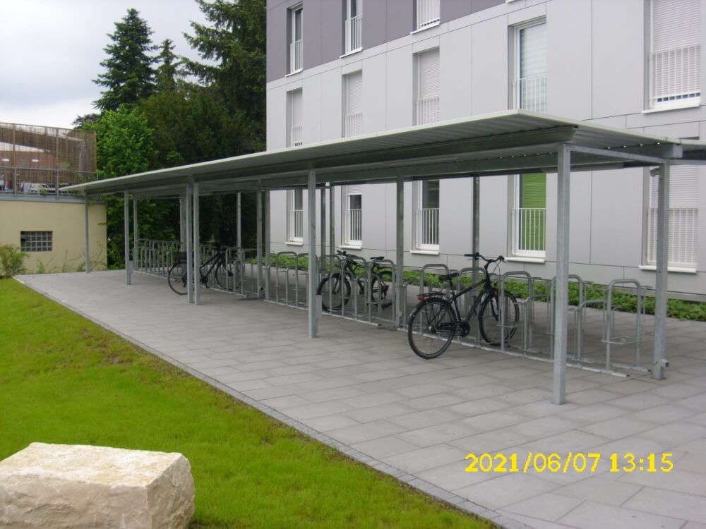 Mehrere Fahrräder stehen in einem überdachten Fahrradständer vor einem modernen Gebäude, in dessen Nähe sich ein Gehweg und Grünflächen befinden. Das Foto trägt unten rechts den Zeitstempel 07.06.2021, 13:15 Uhr.