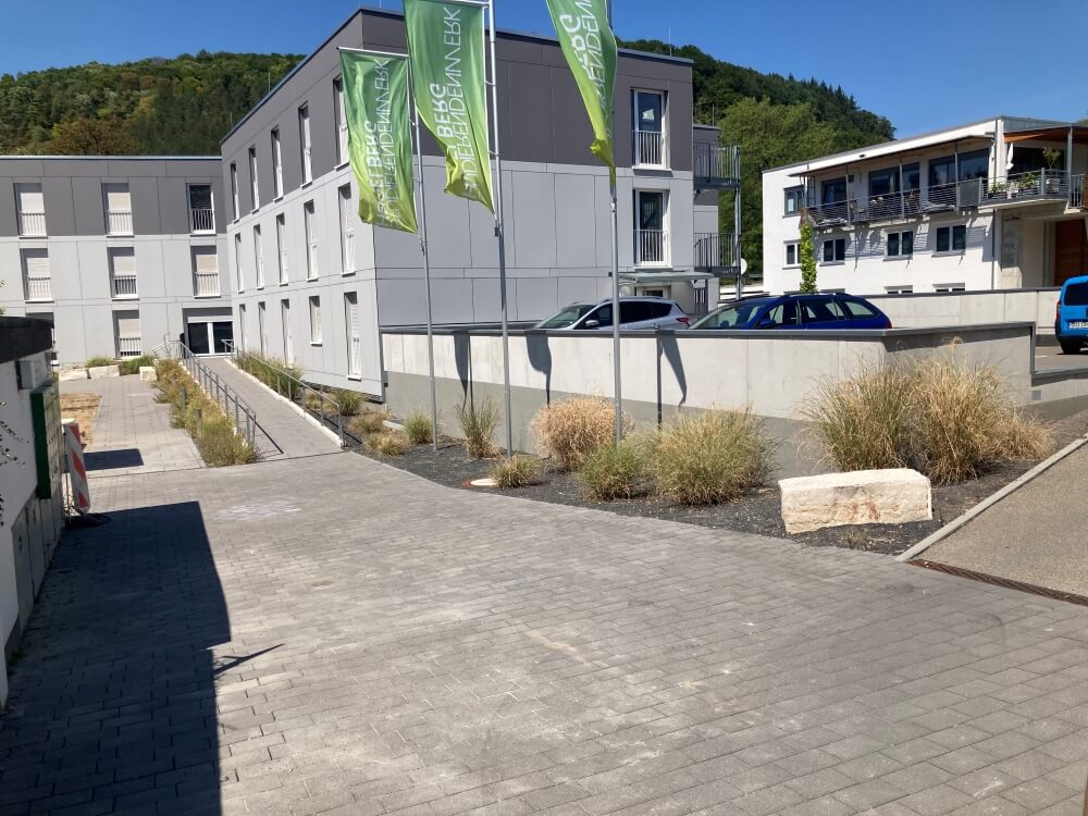 Ein gepflasterter Gehweg führt an modernen weißen Gebäuden mit grünen Fahnen, geparkten Autos und Grünflächen mit hohem Gras vorbei, vor der Kulisse grüner, baumbedeckter Hügel.