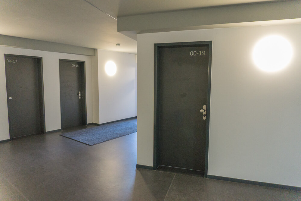 Ein sauberer, moderner Flur mit dunklem Boden und drei dunklen Türen, jede mit weißen Zahlen beschriftet (00-17, 00-18, 00-19). Rechts neben der Tür 00-19 befindet sich eine helle, runde Wandleuchte, im Flur liegt ein blauer Teppich.