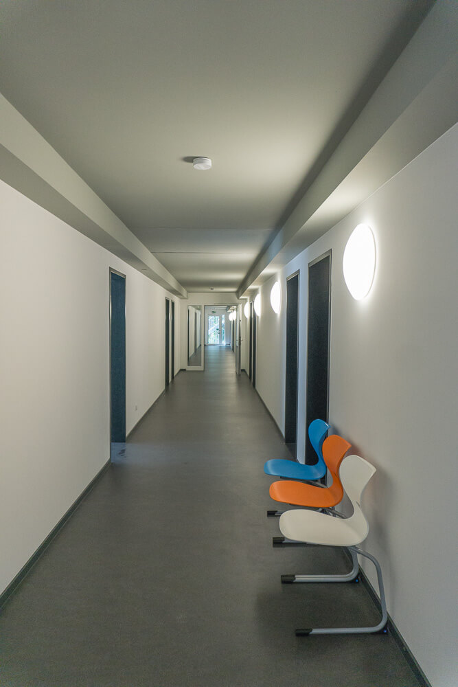 Ein langer, gut beleuchteter Flur mit weißen Wänden und mehreren Türen; drei bunte Stühle (blau, orange und weiß) stehen an der rechten Wand neben einer runden Wandleuchte.