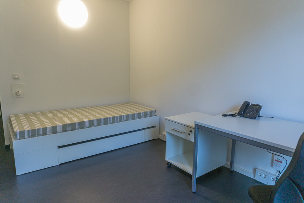 Ein schlichtes, minimalistisches Zimmer mit einem Einzelbett, einem kleinen Schreibtisch mit Telefon, einem Stuhl und einer hellen, runden Wandleuchte. Die Wände sind weiß, der Raum wirkt aufgeräumt und übersichtlich.