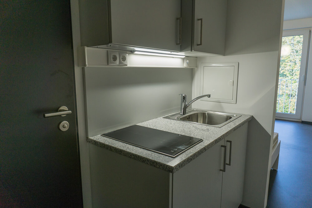 Eine kleine, moderne Küchenzeile mit Spüle, Elektroherd, grauen Schränken und Granitarbeitsplatte. Der Bereich ist gut beleuchtet, im Hintergrund sind eine Tür und ein Fenster zu sehen.