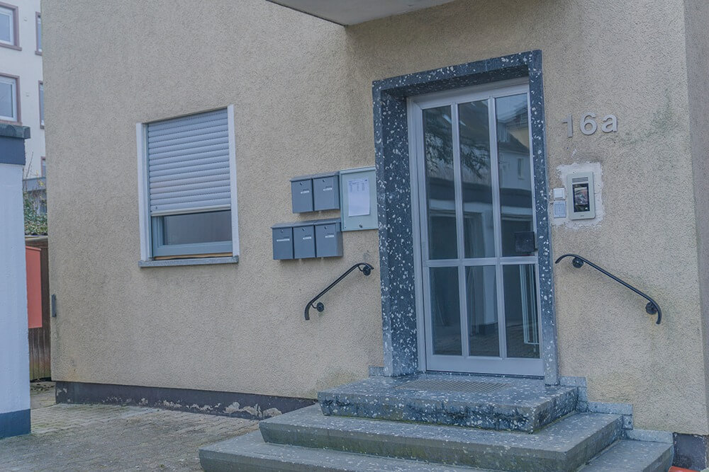 Eingang zu einem beigen Studentenwohnheim, mit einer Glastür, fünf grauen Briefkästen, einem geschlossenen Fenster mit Rollladen, der Nummer 16a an der Wand und zwei schwarzen Handläufen neben Steinstufen.