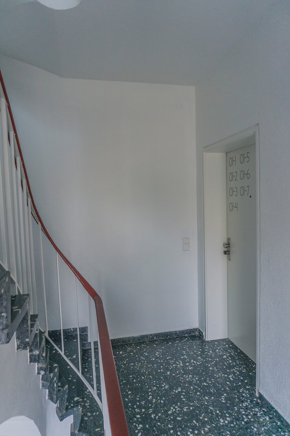 Ein Treppenhaus mit dunklem Terrazzoboden, weißen Wänden und rotem Handlauf. Rechts befindet sich eine weiße Tür mit grauen Zahlen und Buchstaben. Natürliches Licht erhellt den Raum.