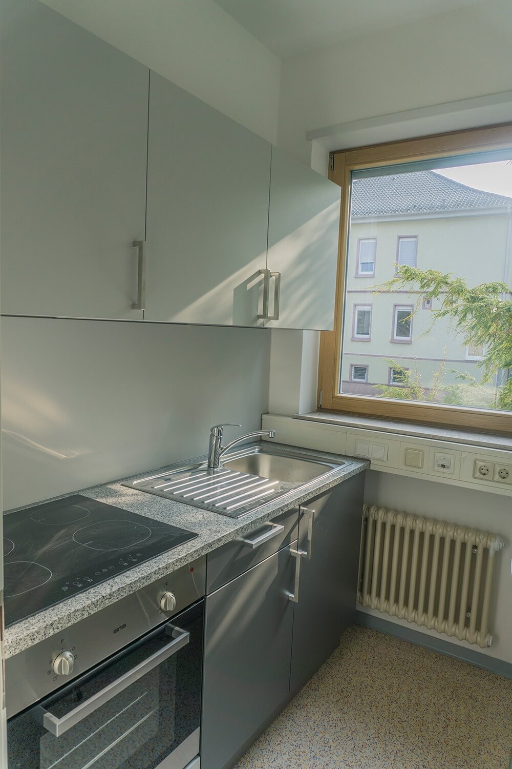 Eine moderne Küche mit grauen Schränken, einem Elektroherd, einer Edelstahlspüle und einem großen Fenster, das natürliches Licht hereinlässt und den Blick auf ein Nachbargebäude und das Grün draußen freigibt.