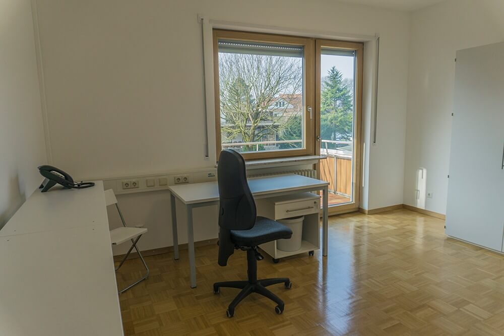 Ein spärlich möbliertes Zimmer mit Schreibtisch, Bürostuhl, Telefon, weißen Schränken und einem Fenster mit Blick auf Bäume und Häuser draußen. Der Raum hat helle Wände und einen Holzboden.