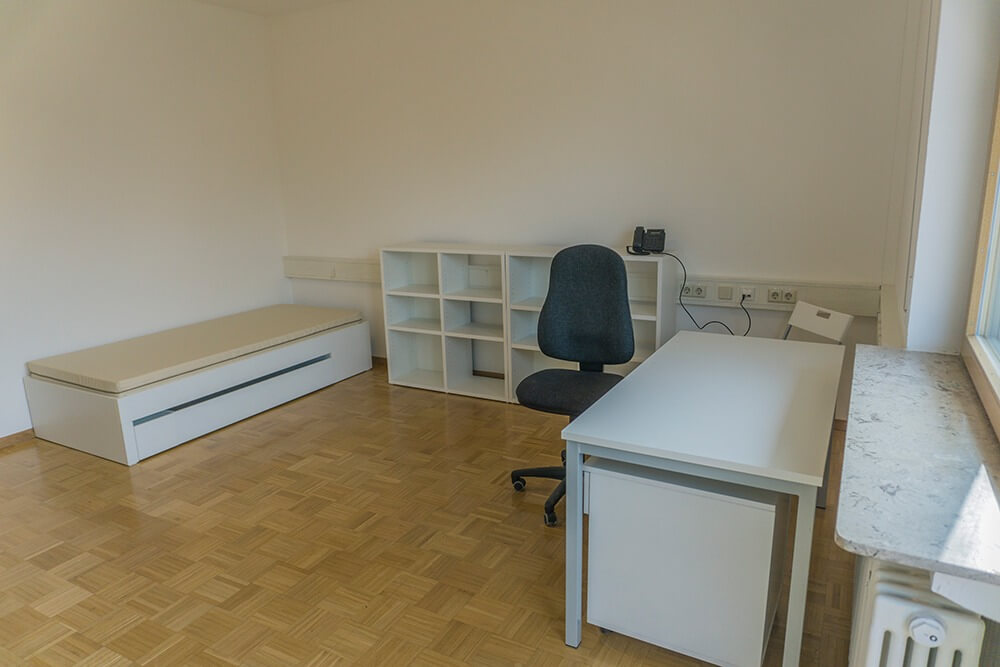 Ein spärlich möbliertes Zimmer mit einem Einzelbett, einem Regal, einem schwarzen Bürostuhl und einem weißen Schreibtisch auf hellem Holzboden. Der Raum hat schlichte weiße Wände und ein Fenster auf der rechten Seite.