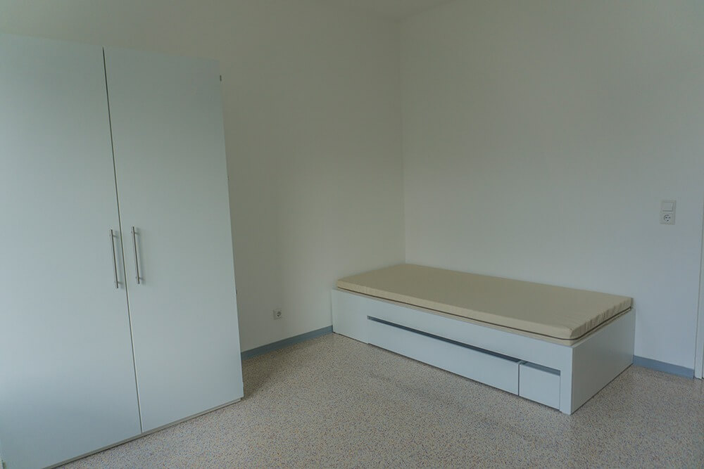 Ein minimalistisches Zimmer mit einem geschlossenen weißen Kleiderschrank auf der linken Seite und einem einzelnen weißen Bett mit einer beigen Matratze auf der rechten Seite, vor schlichten weißen Wänden und einem gesprenkelten beigen Boden.