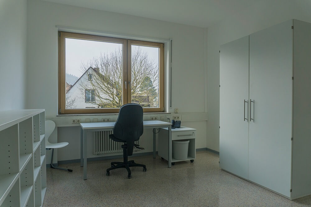 Ein minimalistisches Zimmer mit weißem Schreibtisch, schwarzem Stuhl, weißen Regalen, einem Schrank und einem Fenster mit Blick auf Bäume und Häuser an einem bewölkten Tag. Der Raum ist ordentlich und gut beleuchtet.