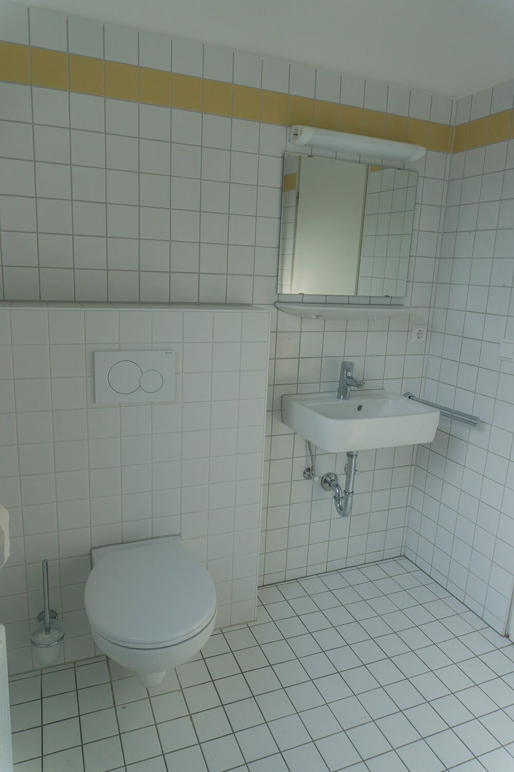 Ein sauberes, weiß gefliestes Badezimmer mit einer wandmontierten Toilette, einem Toilettenbürstenhalter, einem rechteckigen Waschbecken mit einem Spiegel darüber und einem gelben Streifen an der Decke.