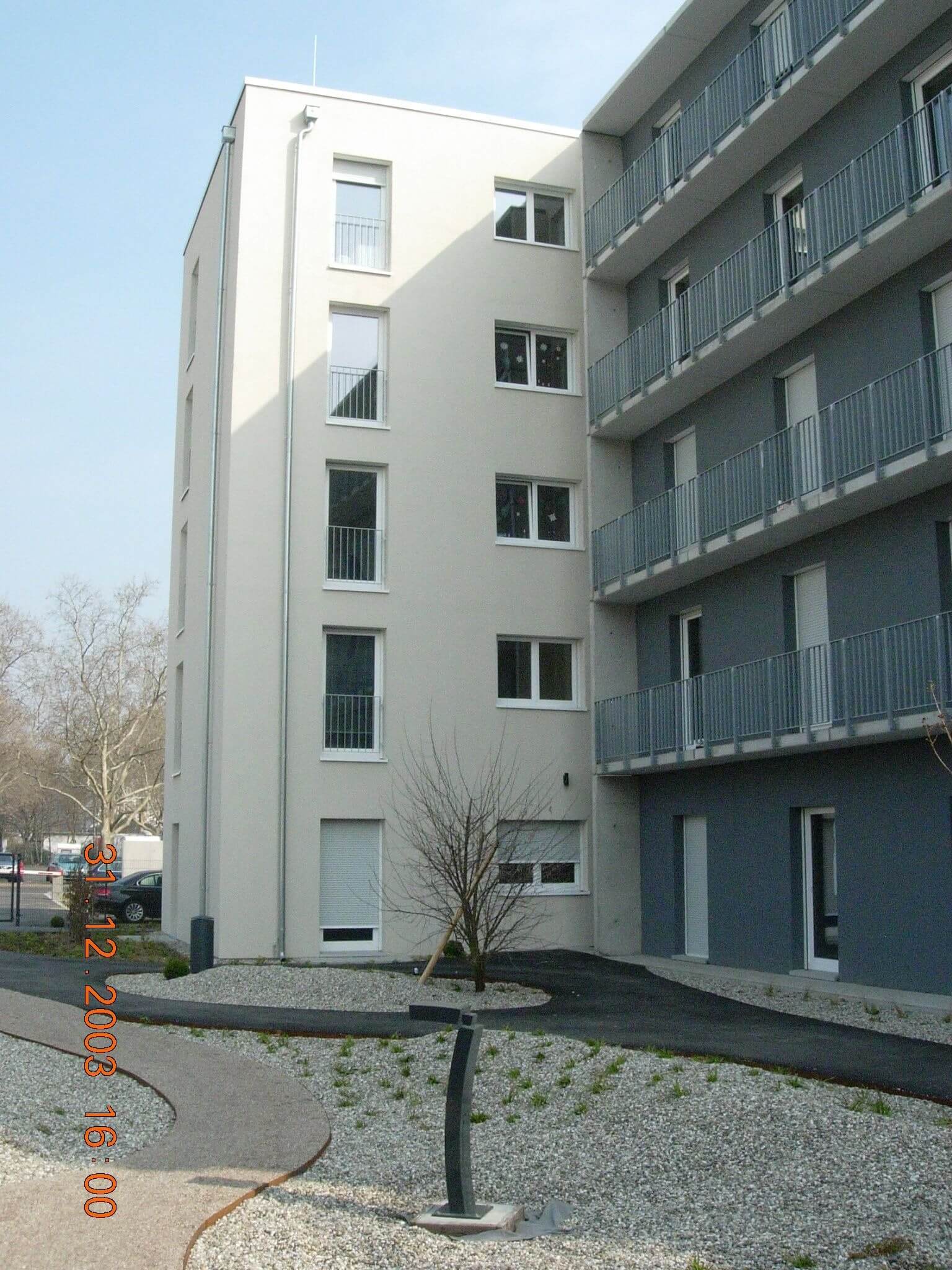 Seitenansicht eines hellen Wohngebäudes mit grauem Anbau, von Wegen umgeben, die durch Kiesbeete mit junger Bepflanzung führen.