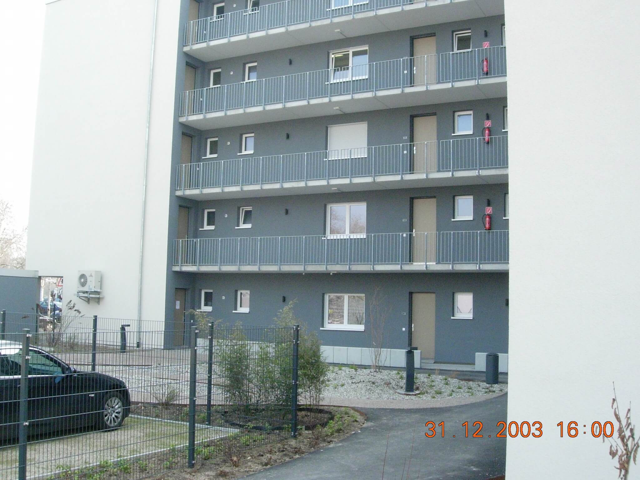 Rückansicht eines modernen Wohnhauses mit mehreren Balkonen und Wohnungseingängen, im Vordergrund ein kleiner Innenhof mit Kiesflächen und Pflanzbeeten.