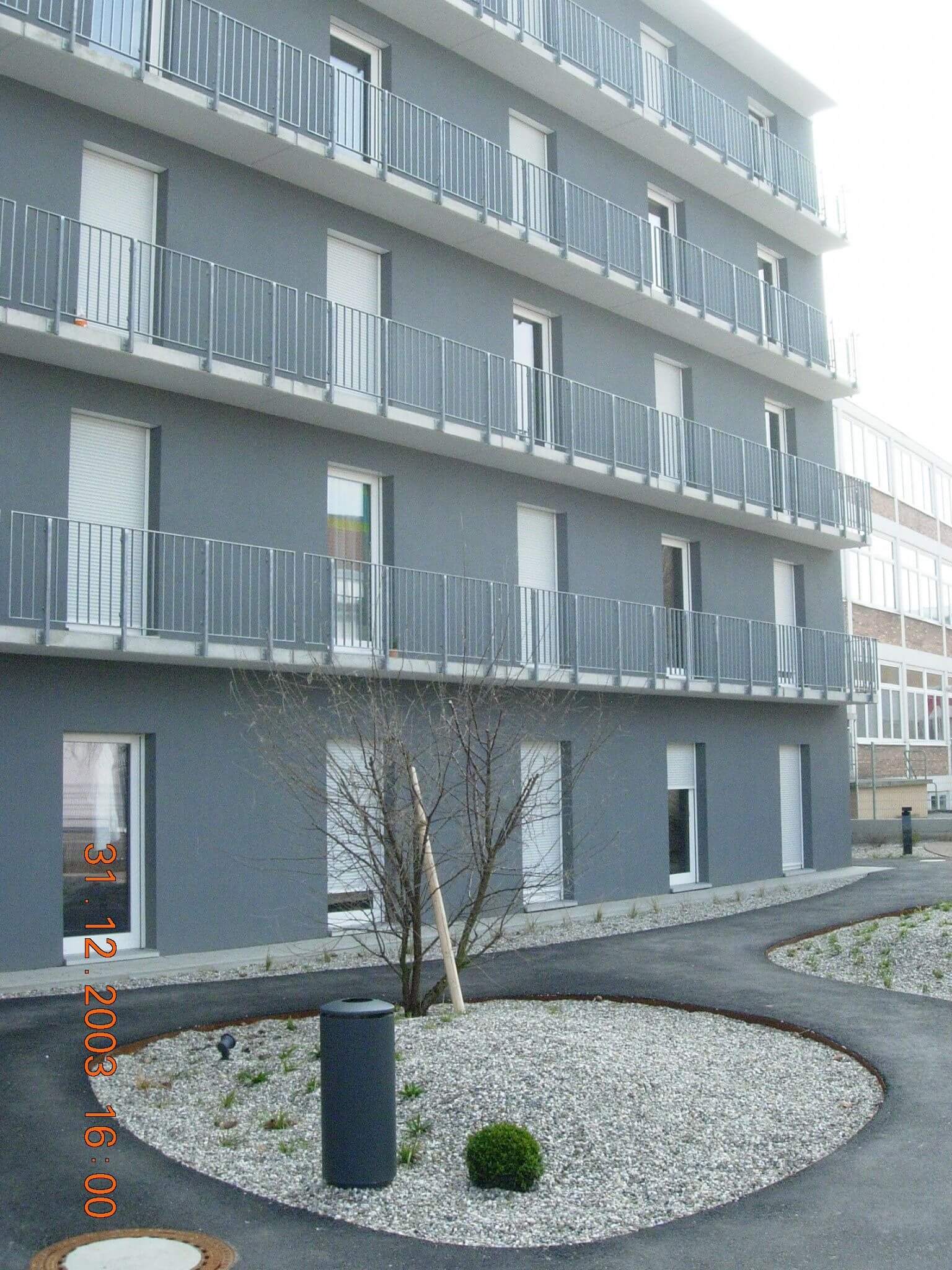 Mehrstöckiges Wohngebäude mit grauer Fassade und weißen Fenstern, vorgelagertem Kiesbeet und Balkonen mit Metallgeländern.
