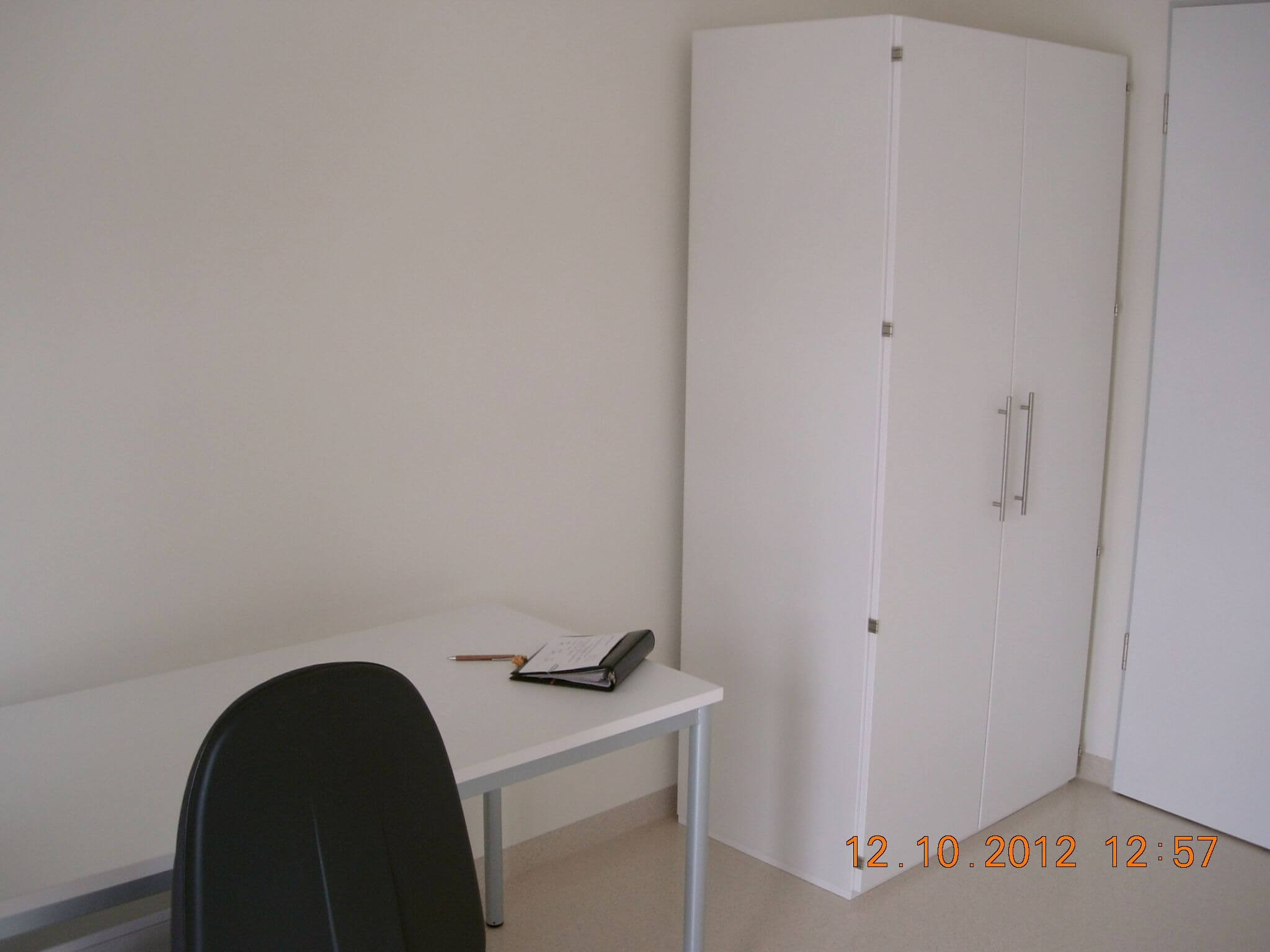 Ein einfacher Arbeitsplatz mit weißem Schreibtisch, schwarzem Stuhl und einem weißen Kleiderschrank in einer hellen Zimmerecke.