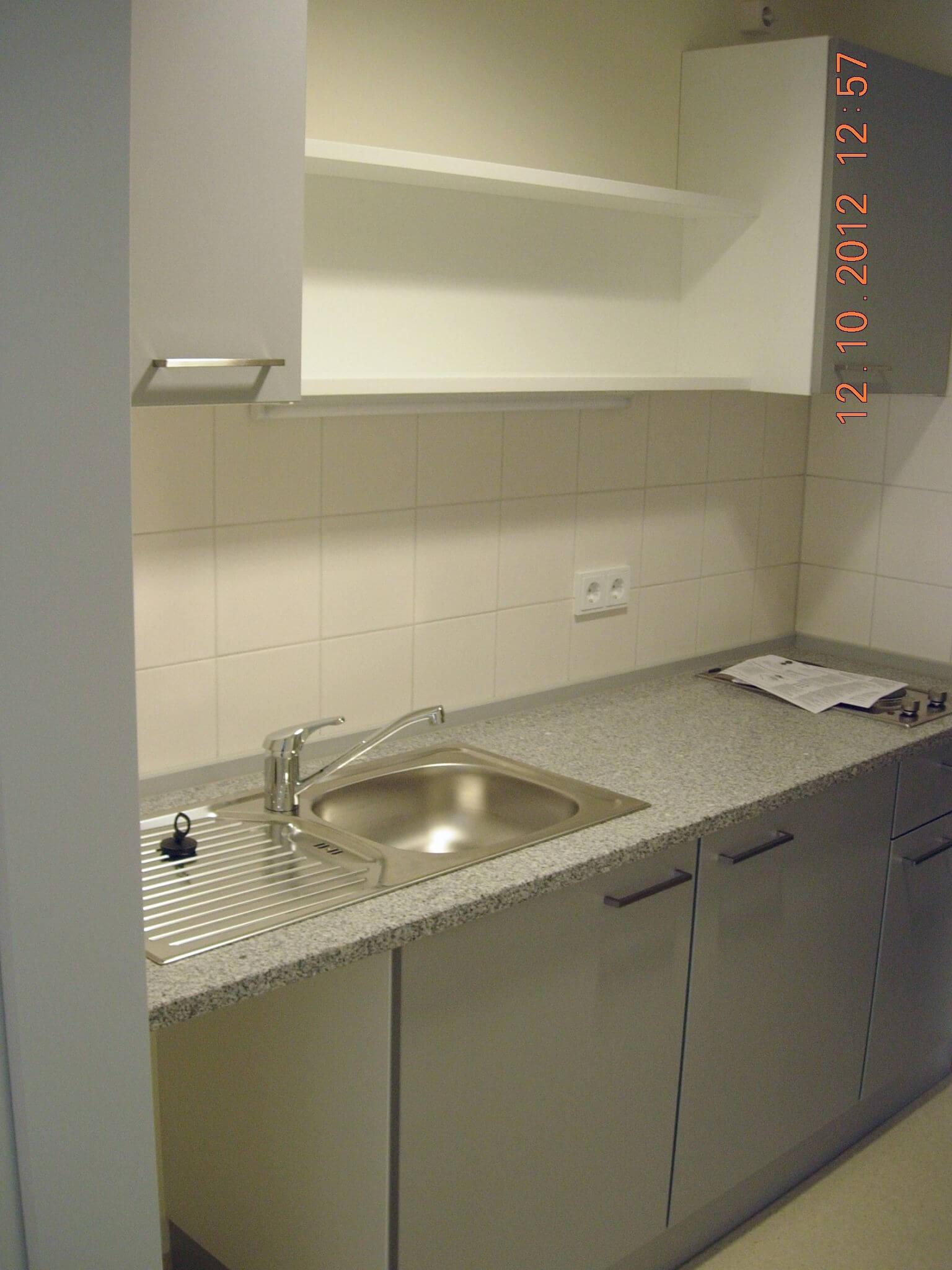 Moderne Küchenzeile mit grauer Arbeitsfläche, Edelstahlspüle und weißen Oberschränken vor weiß gefliester Wand.