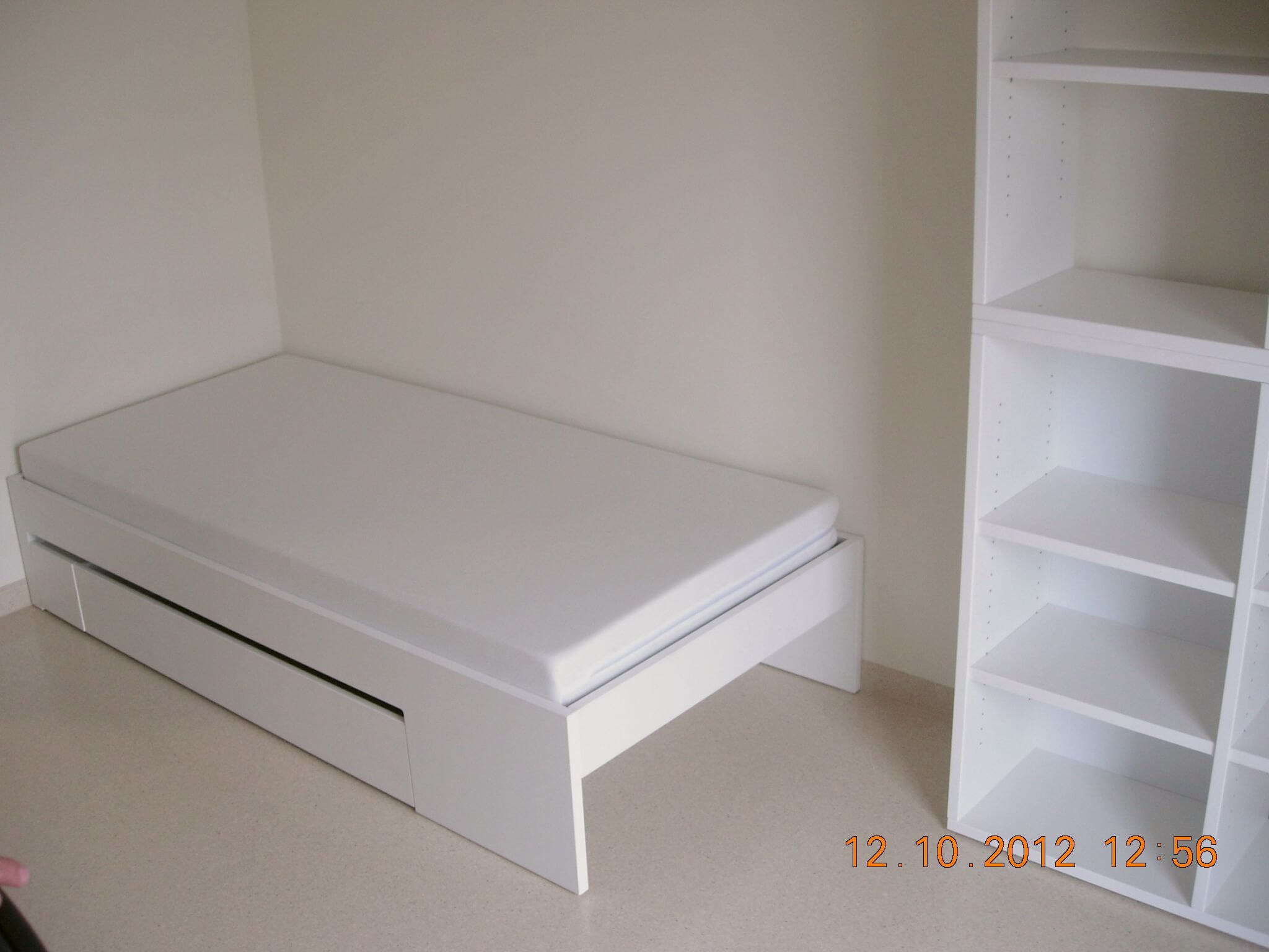Ein schlicht eingerichtetes Zimmer mit einem weißen Einzelbett und einem weißen Regal an der Wand, beide Möbelstücke minimalistisch gestaltet.