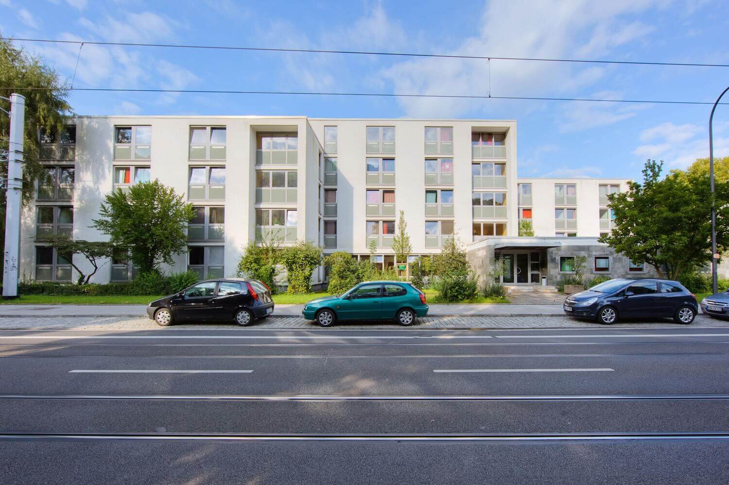 Moderne Außenansicht eines mehrstöckigen Studentenwohnheims mit großen Fenstern, Vorgarten, parkenden Autos und einer mehrspurigen Straße mit Straßenbahnschienen im Vordergrund