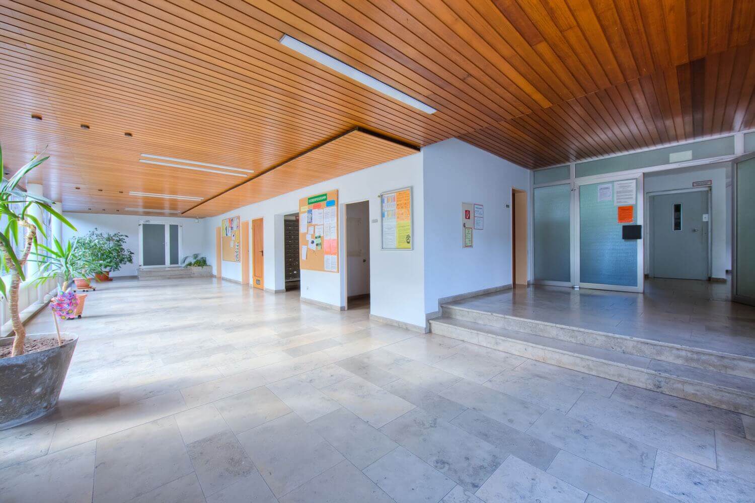 Weitläufiges, helles Foyer mit Pflanzen, Pinnwänden und Holzelementen an Decke und Türen in einem Studentenwohnheim