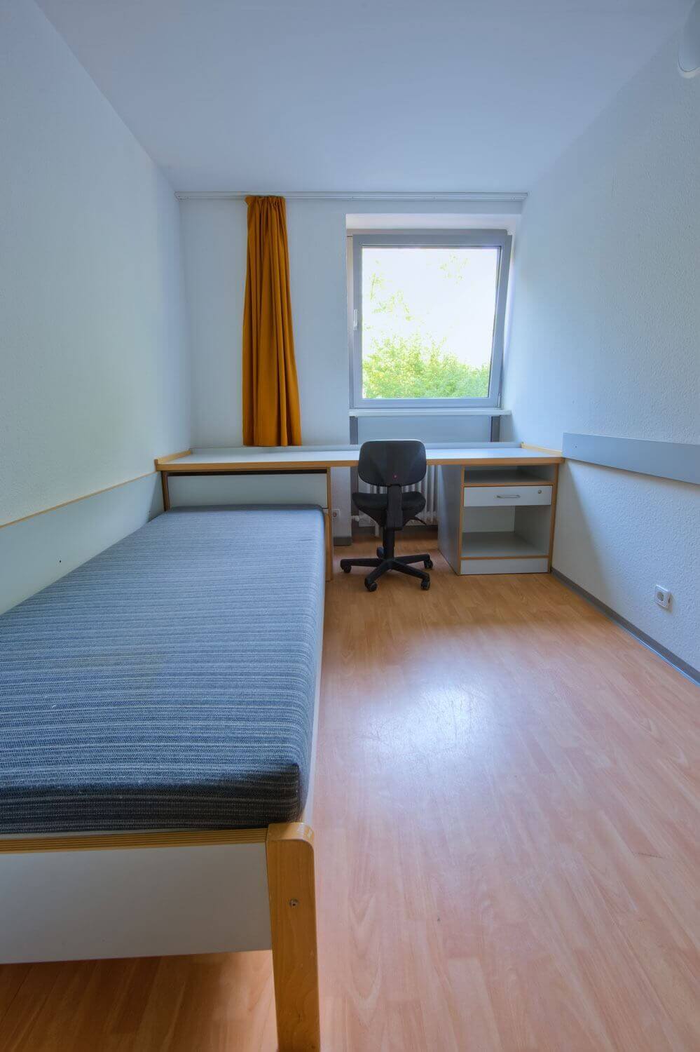 Einzelzimmer mit Bett, großem Fenster, langem Schreibtisch und Drehstuhl in einem Studentenwohnheim