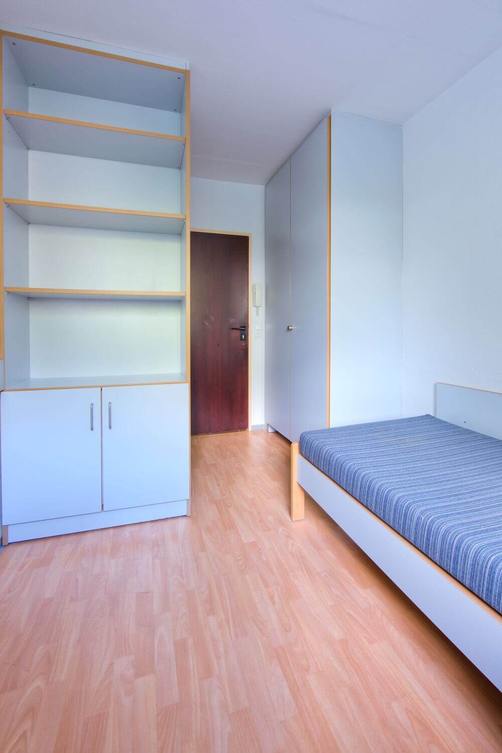 Kompakt eingerichtetes Einzelzimmer mit Bett, Kleiderschrank, Regal und Schrankelement nahe der Zimmertür