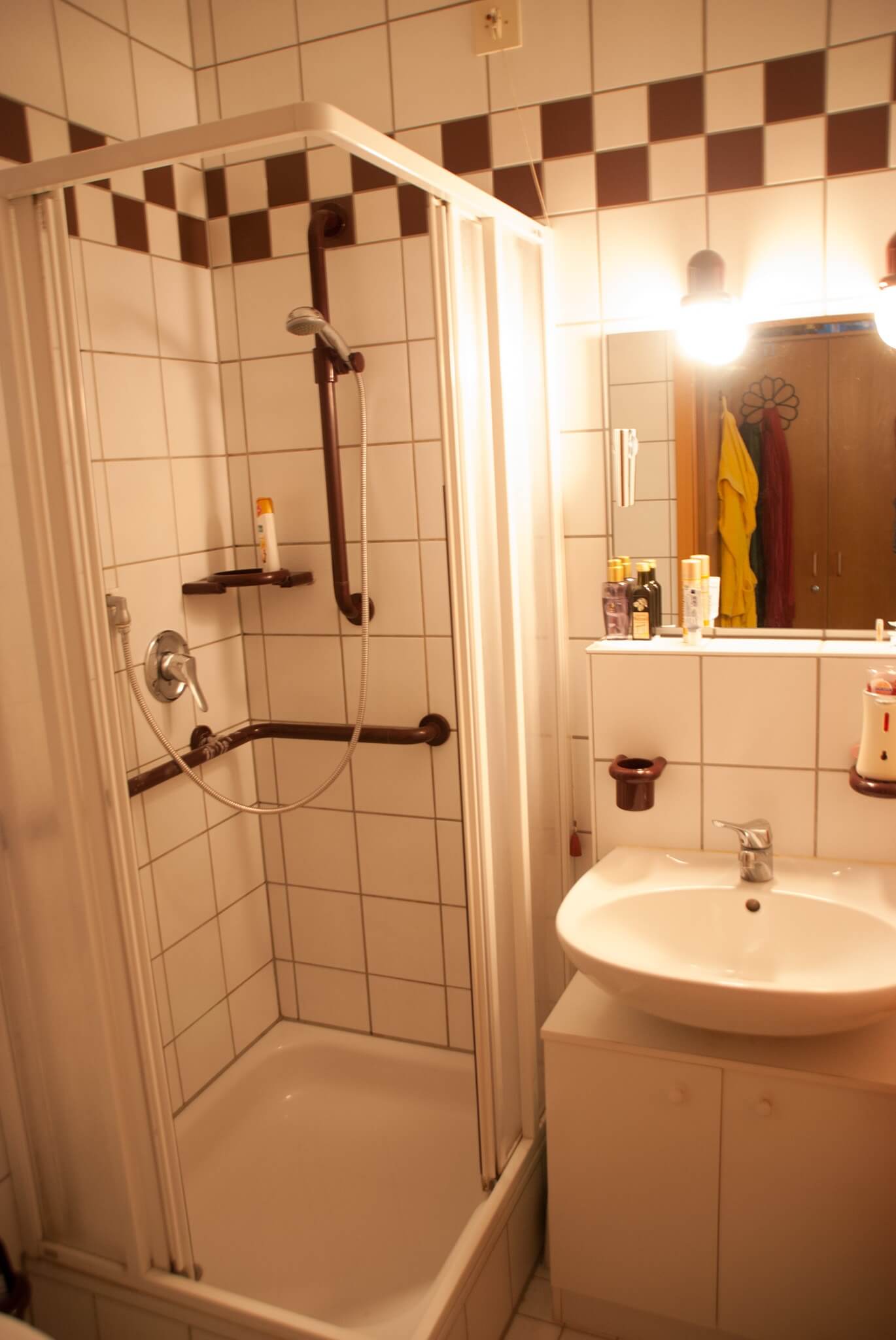 Ein Badezimmer mit weiß gefliester Dusche, Haltegriffen und Waschbecken. Auf dem Spiegelregal stehen verschiedene Pflegeprodukte, das Licht ist warm.