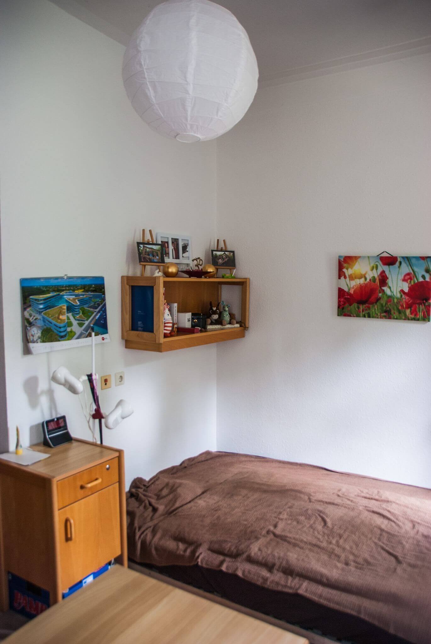 Ein gemütlich dekoriertes Einzelzimmer mit brauner Bettdecke, Wandbildern und einem Regal mit persönlichen Gegenständen. Eine weiße Papierlampe hängt von der Decke.