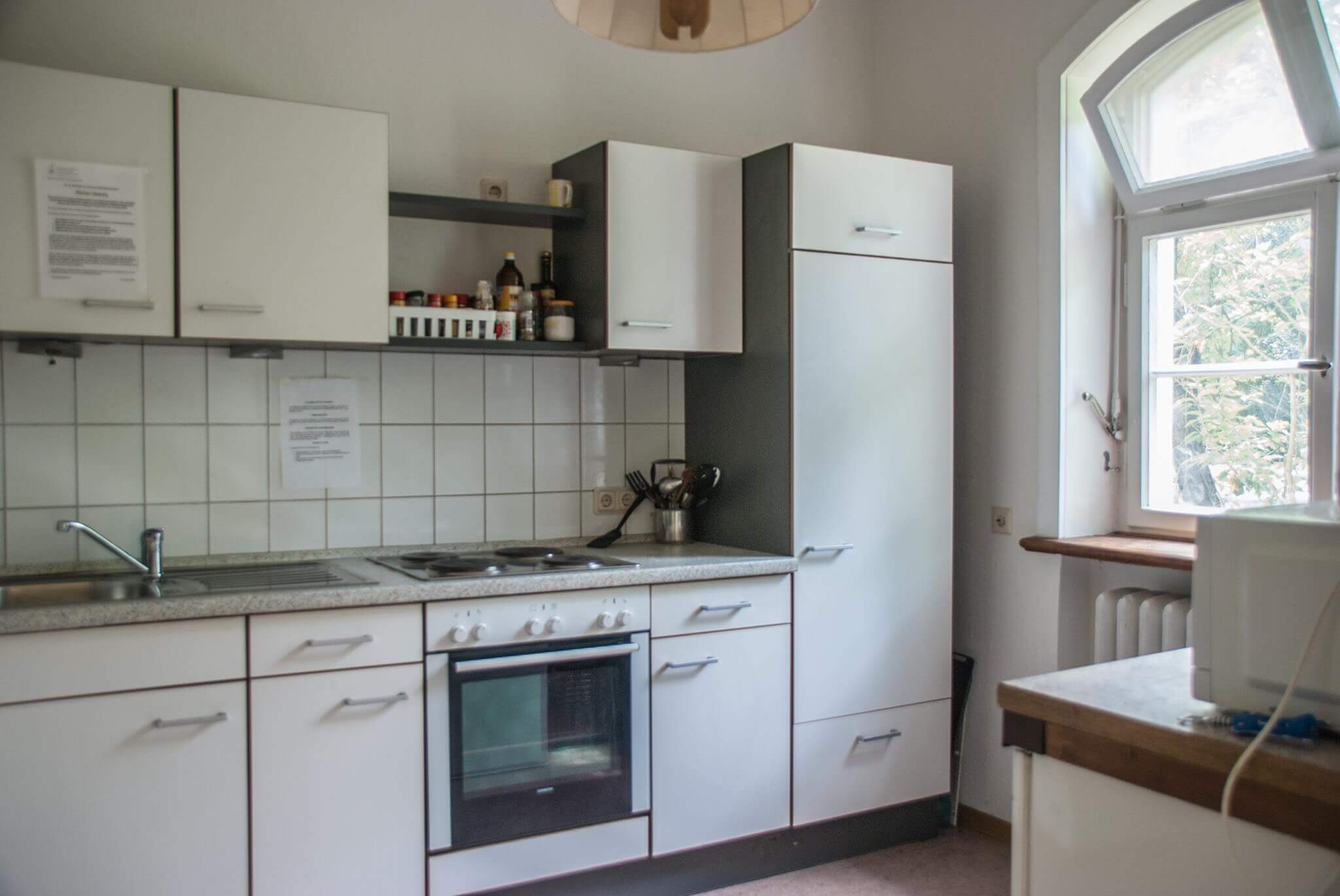 Eine kompakte Küche mit weißer Einbauzeile, Herd und Spüle. Auf dem Schrank neben der Fensterbank steht eine Mikrowelle, im Regal sind Gewürze und Küchengeräte zu sehen.