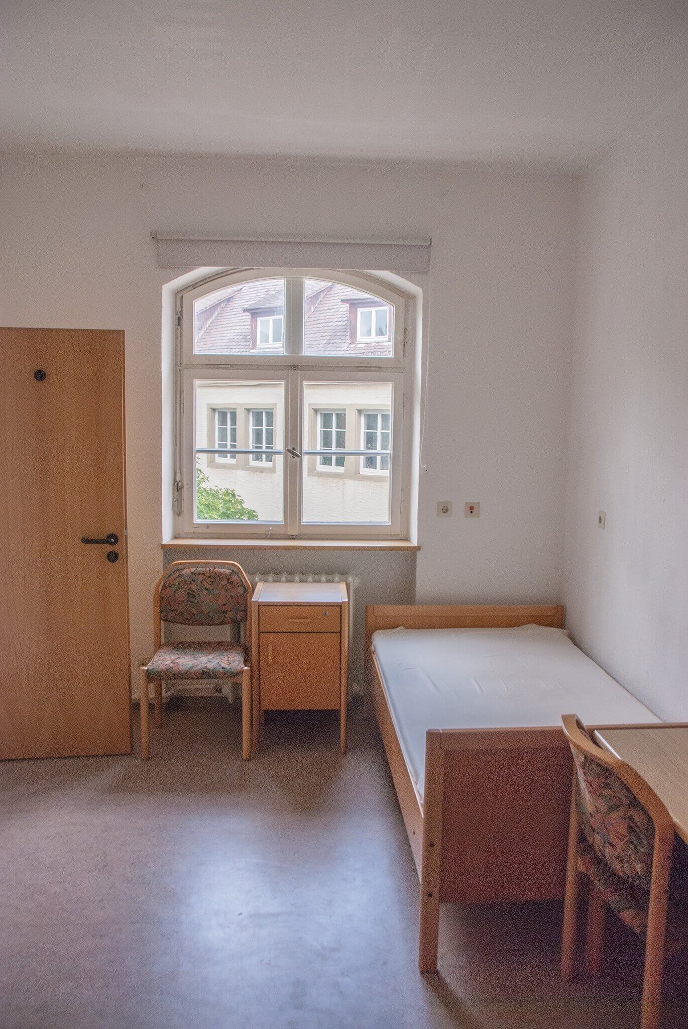 Ein schlicht eingerichtetes Einzelzimmer mit Bett, Nachttisch und zwei Stühlen. Durch das Fenster fällt Tageslicht auf die weißen Wände und den braunen Bodenbelag.