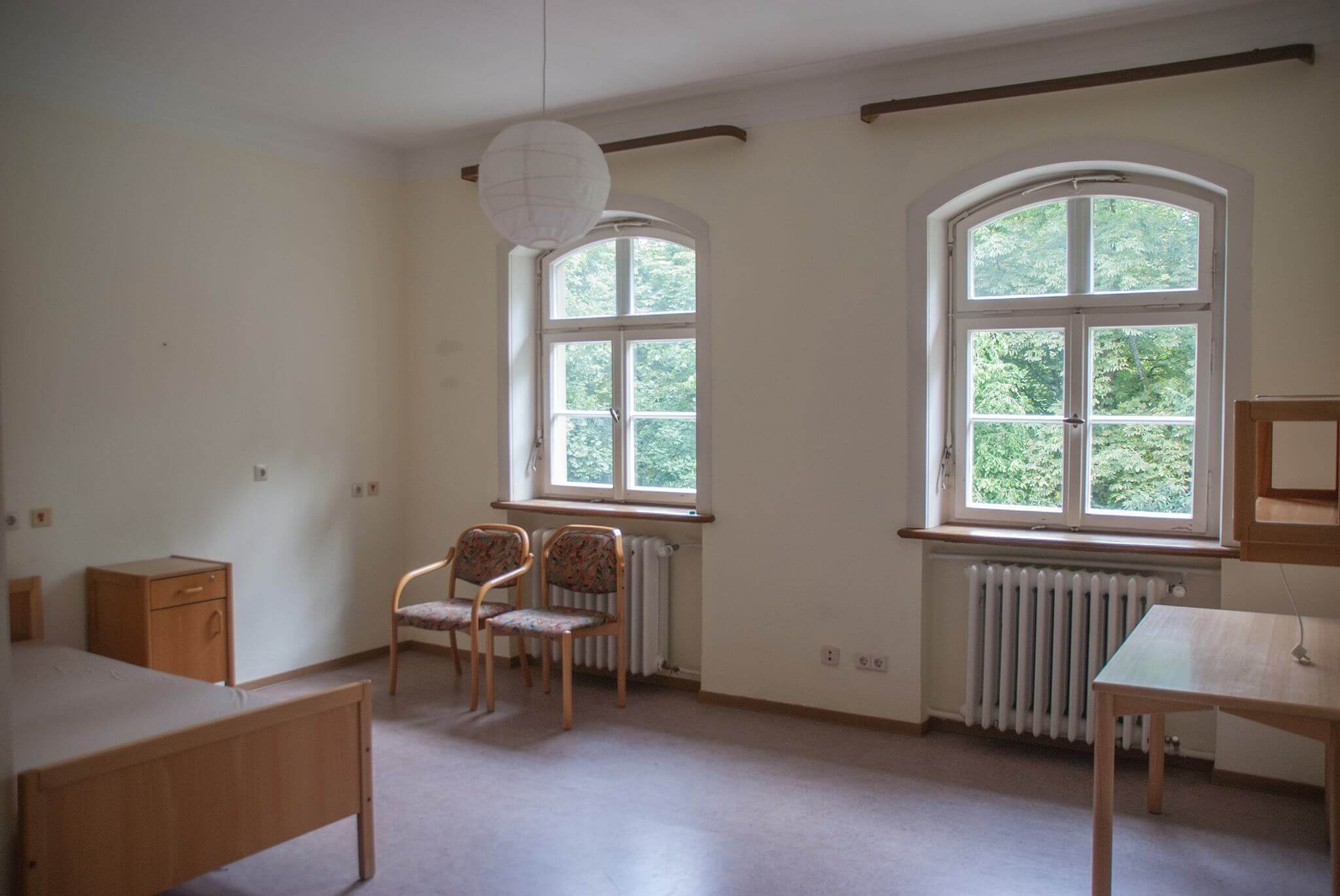 Ein helles Zimmer mit zwei großen Fenstern, zwei bunten Holzstühlen und einfacher Möblierung, darunter ein Bett, ein Tisch und ein Nachtschrank. Der Raum wirkt aufgeräumt und freundlich.