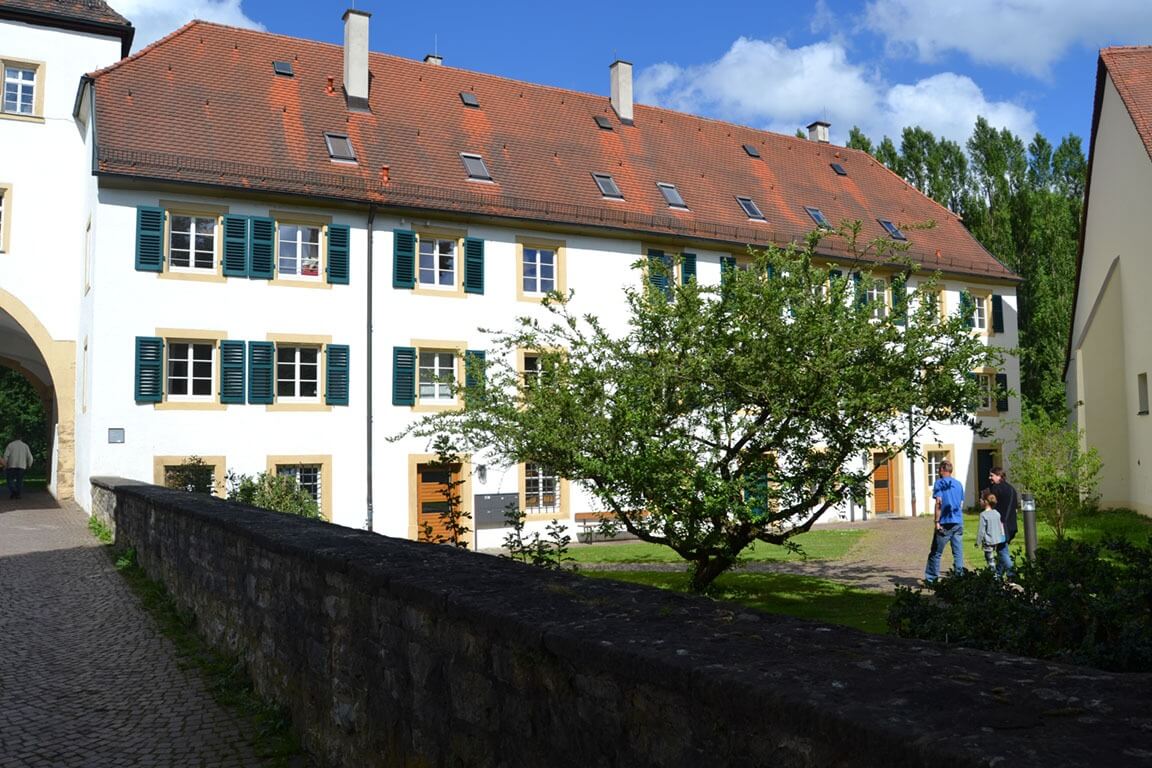 Ein historisches Gebäude mit weißer Fassade, grünen Fensterläden und rotem Dach; davor ein gepflasterter Weg und ein kleiner Baum im Innenhof.