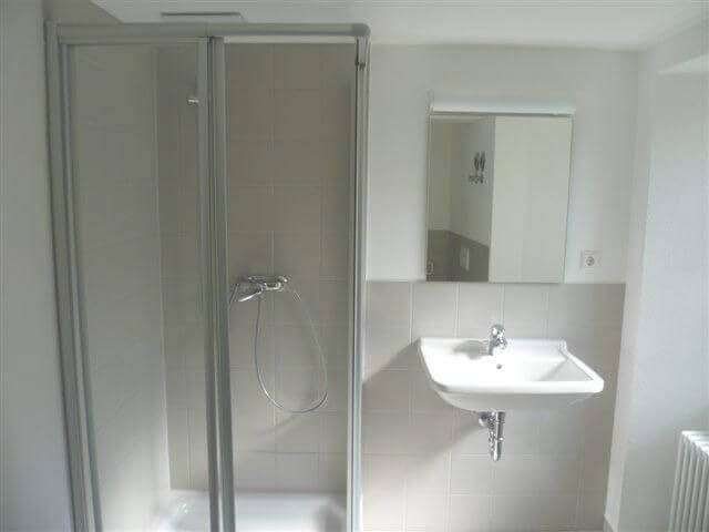 Ein schlichtes Badezimmer mit gefliester Dusche, weißem Waschbecken, Spiegel und Handtuchheizung an der Wand.