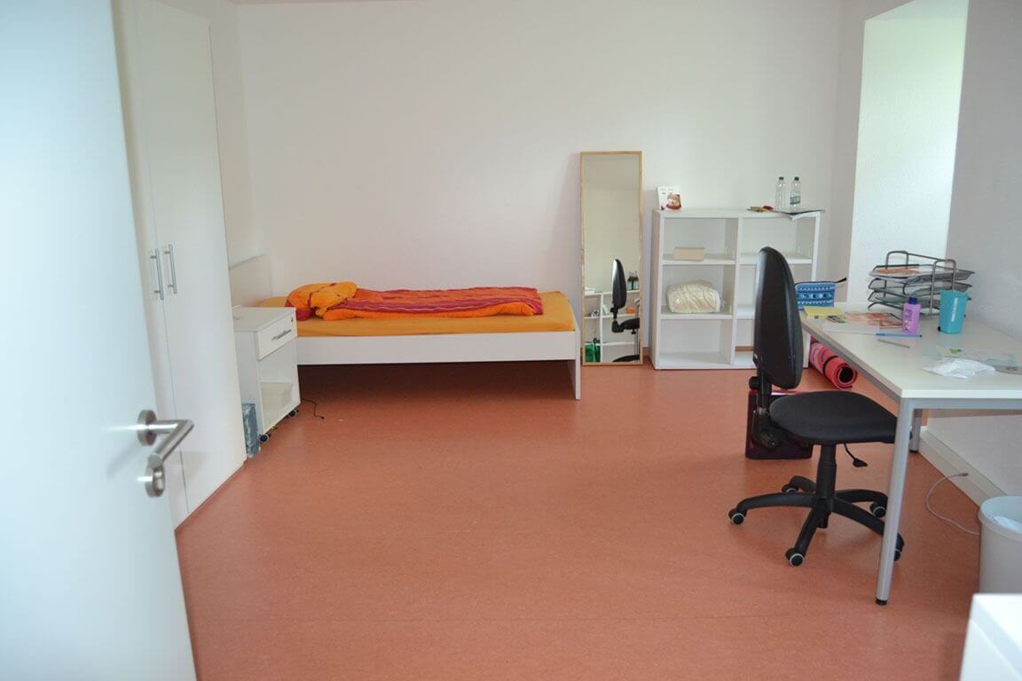 Ein großzügiges Einzelzimmer mit Schreibtisch, Regal, Spiegel und Bett mit orangefarbener Bettwäsche, ausgestattet mit einfachen weißen Möbeln.