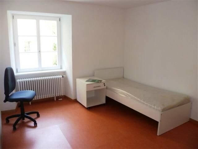 Ein einfach möbliertes Zimmer mit Einzelbett, weißem Nachttisch und Schreibtischstuhl vor einem Fenster mit Blick nach draußen.