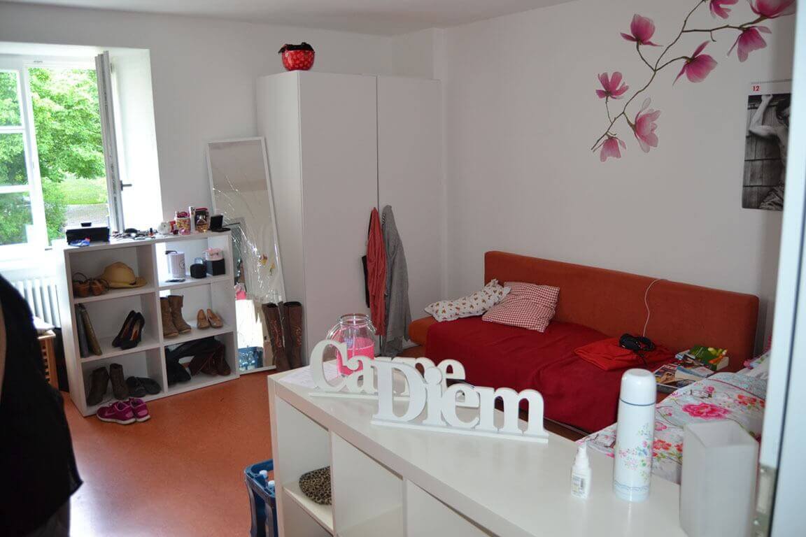 Ein gemütlich eingerichtetes Zimmer mit offenem Fenster, einem roten Sofa, weißen Möbeln und Wanddeko in Form von rosafarbenen Blüten.