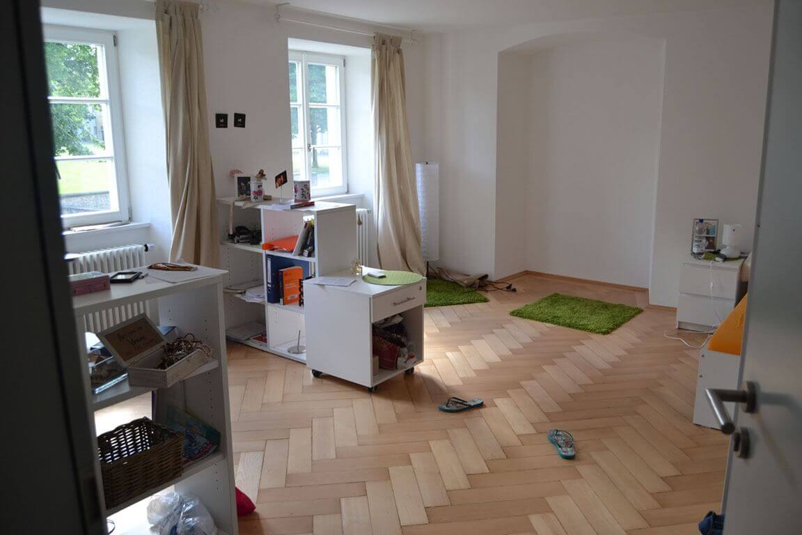 Ein geräumiges, helles Zimmer mit zwei großen Fenstern, hellem Parkettboden und weißen Möbeln; an der Wand hängen helle Vorhänge, auf dem Boden liegen zwei grüne Teppiche.