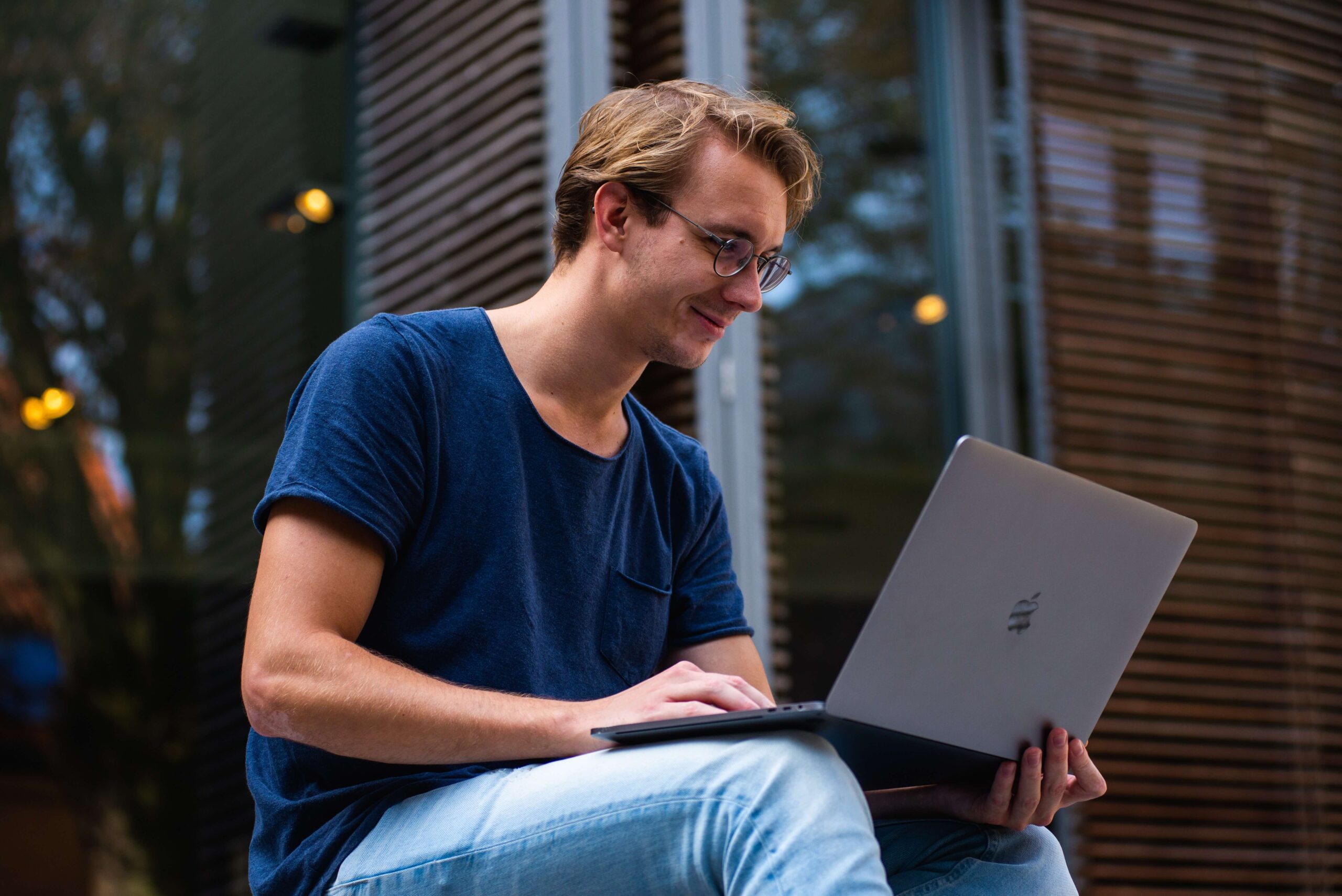 Ein junger Mann sitzt entspannt im Freien auf einer Treppe und arbeitet konzentriert an seinem Laptop. Im Hintergrund spiegeln sich Fensterläden in der Glasfassade.