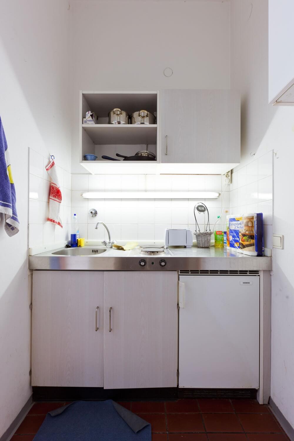 Kompakte Küchenzeile mit Spüle, zwei Kochplatten, Kühlschrank, Hängeschränken und Fliesenspiegel.