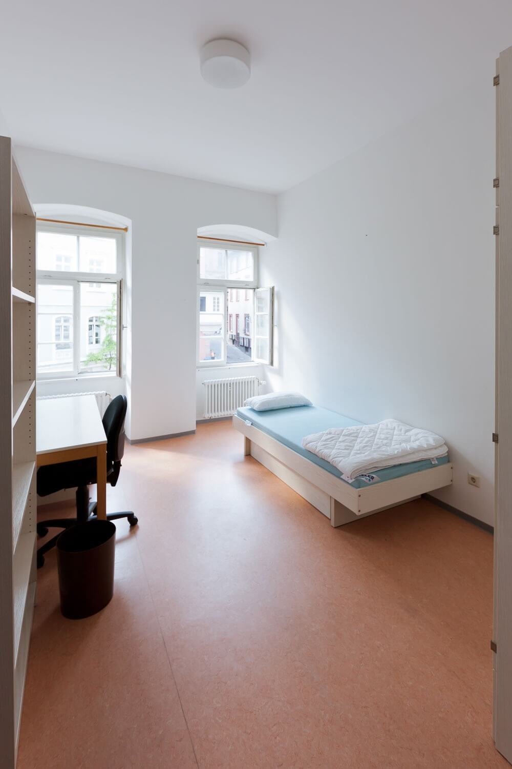 Freundlich eingerichtetes Zimmer mit zwei Fenstern, Schreibtisch, Stuhl, Regal und Einzelbett mit hellblauer Matratze.