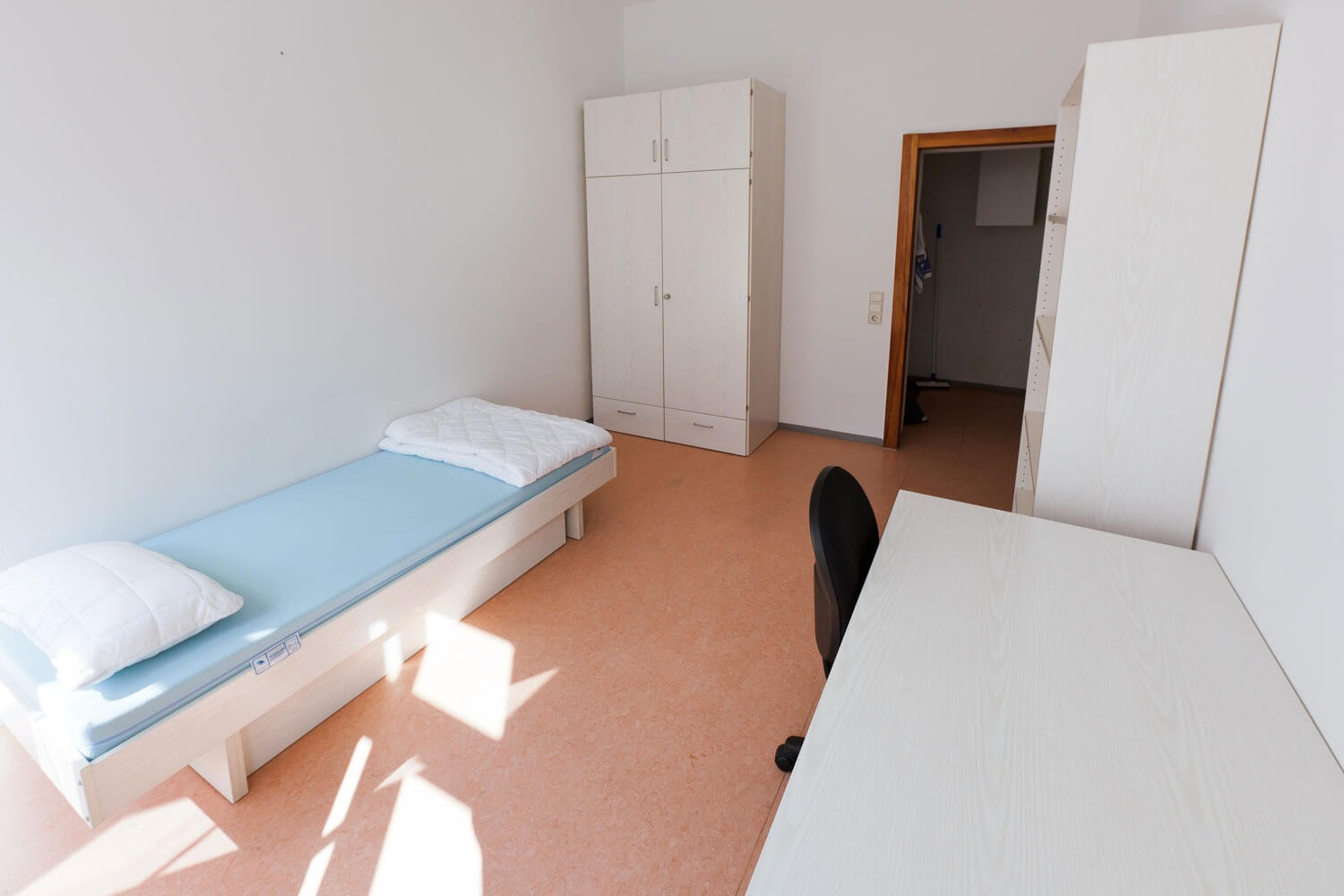 Helles Einzelzimmer mit Bett, Schreibtisch, schwarzem Stuhl, Kleiderschrank und Regal auf hellbraunem Boden.