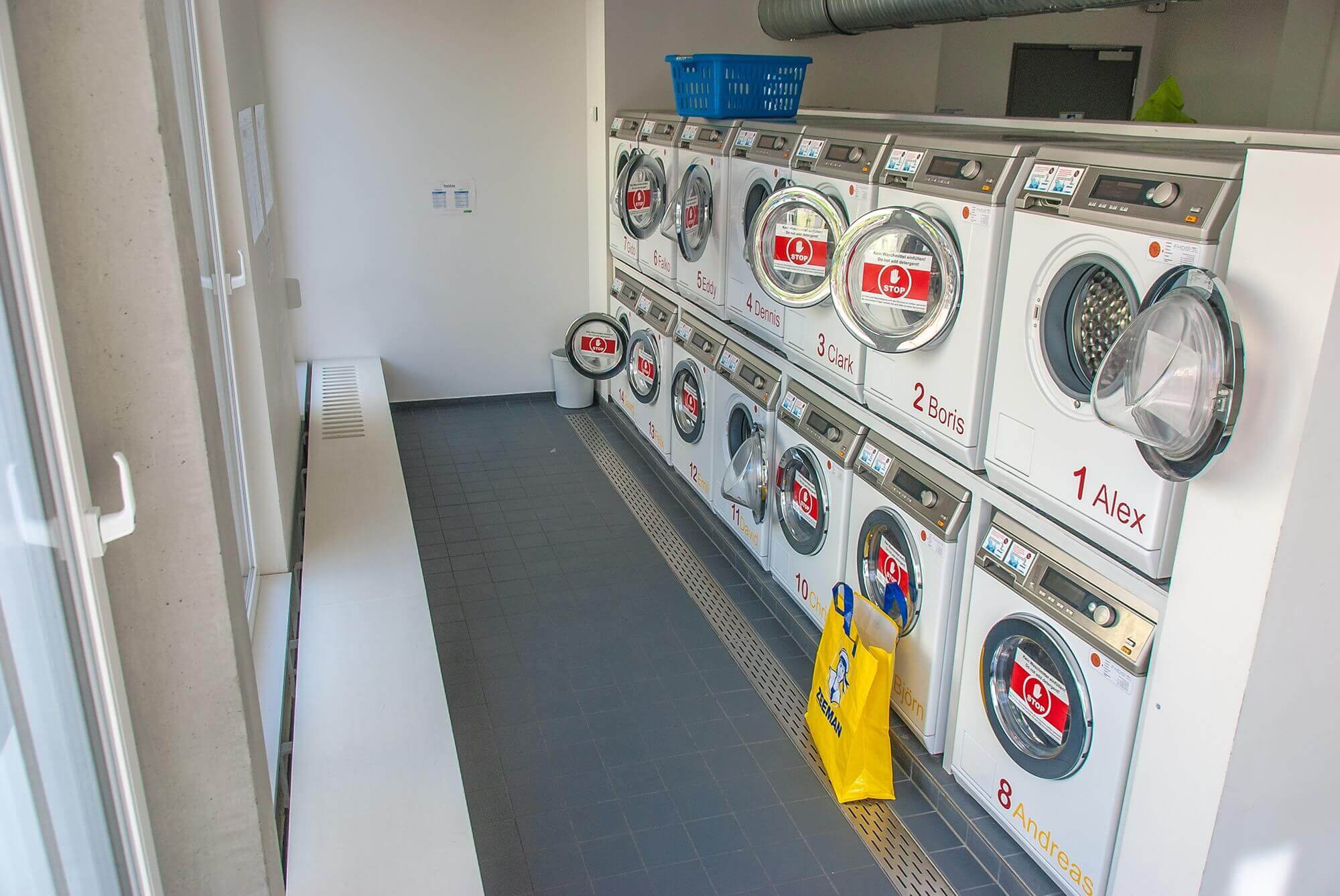 Reihe moderner Waschmaschinen. in einem hellen Waschraum mit grauem Fliesenboden, großen Fenstern auf der linken Seite und einer gelben Einkaufstasche auf dem Boden neben dem Gerät mit der Aufschrift „Alex“.